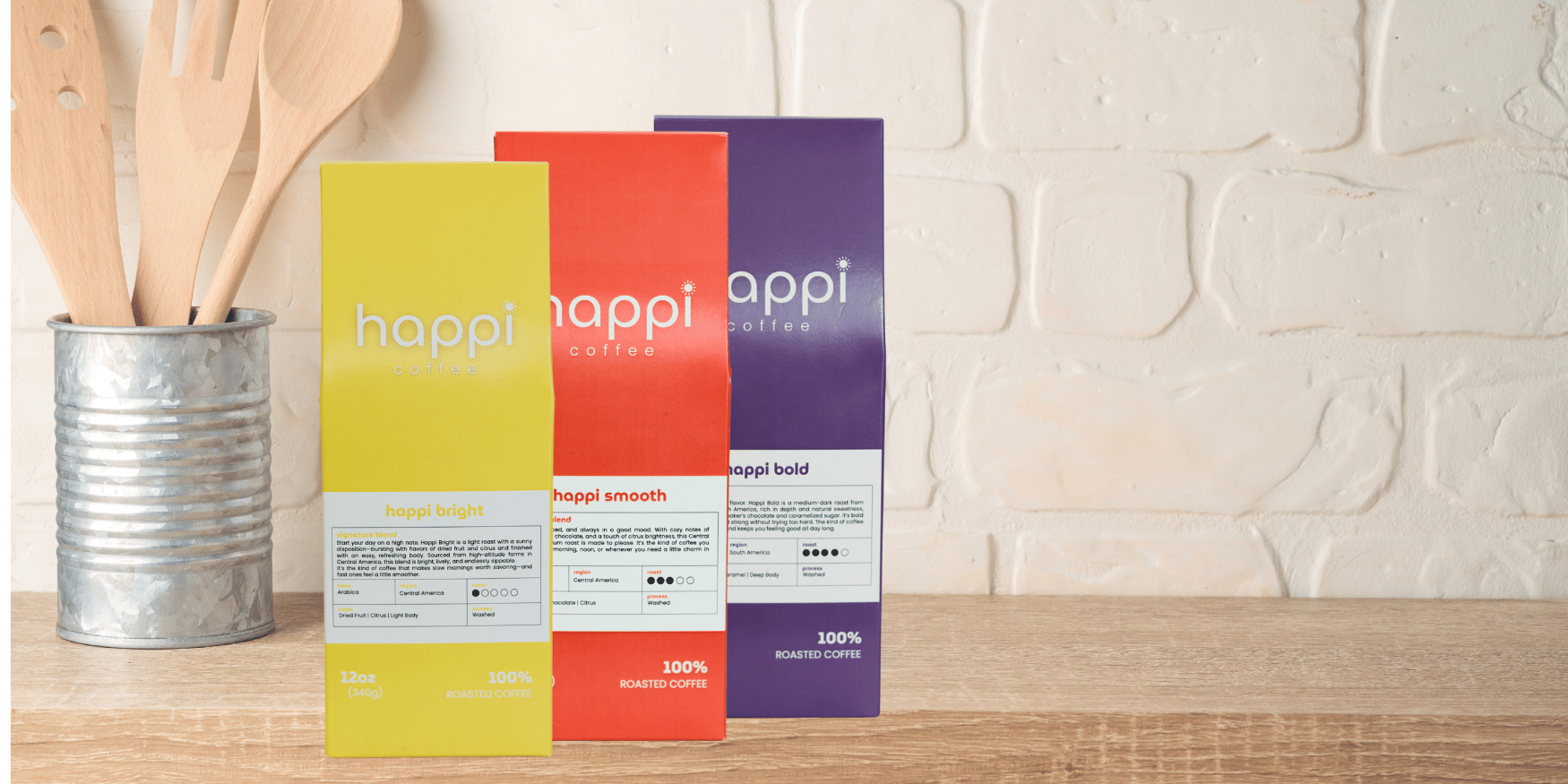 Three colorful bags of Happi Coffee signature blends—Happi Bright (light roast), Happi Smooth (medium roast), and Happi Bold (dark roast)—displayed on a wooden counter next to a metal tin of kitchen utensils.