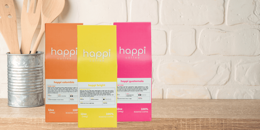 Happi Coffee light roast lineup—Happi Bright, Happi Colombia, and Happi Guatemala bags on a kitchen counter. Freshly roasted Arabica coffee, available in whole bean or ground.