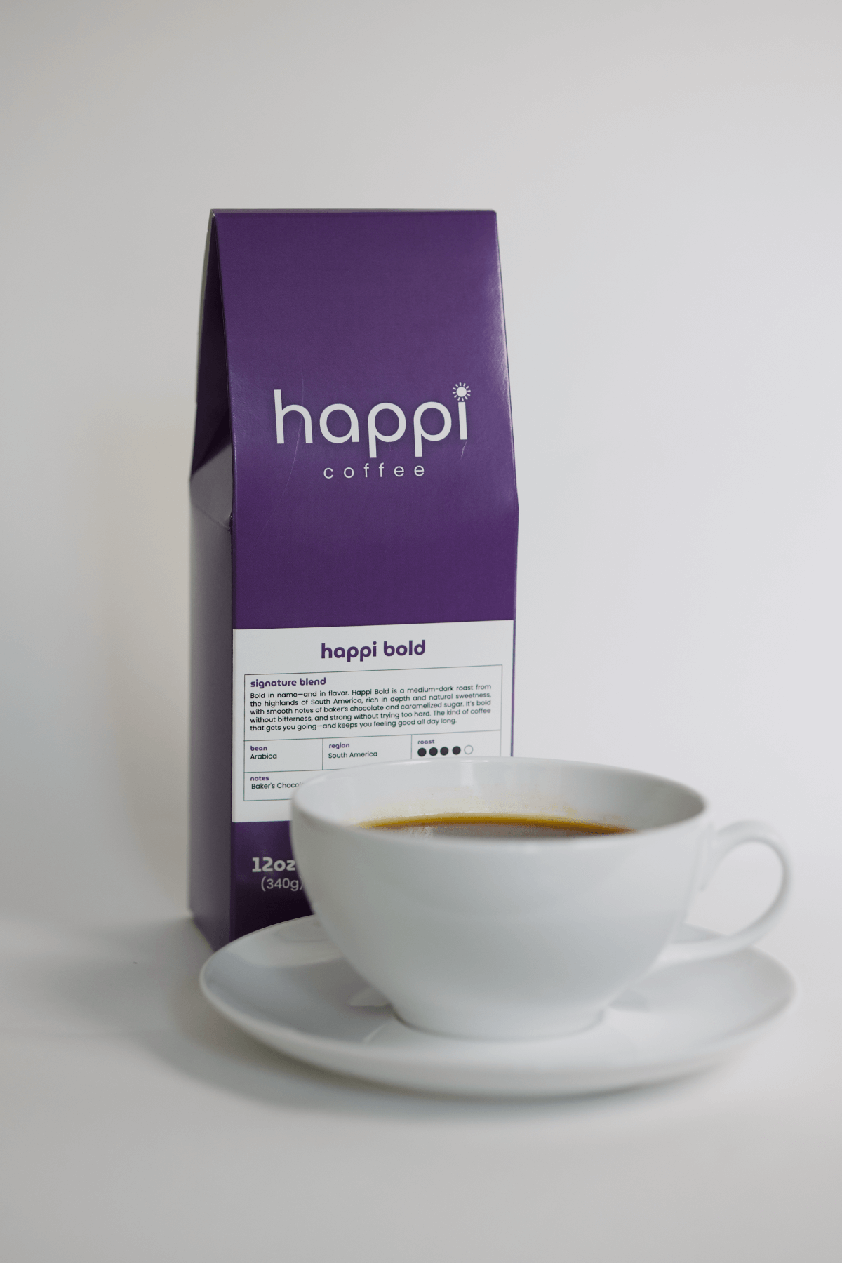 12 oz bag of happi bold dark roast specialty coffee with brewed coffee in a white cup and saucer