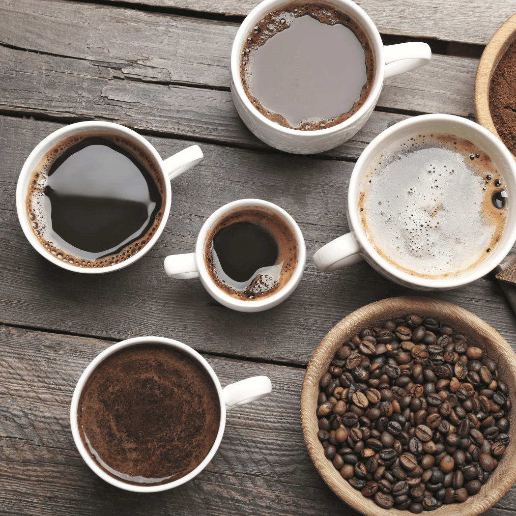 Assorted cups of coffee showing different brew styles, alongside a wooden bowl of roasted coffee beans on a rustic wooden table.