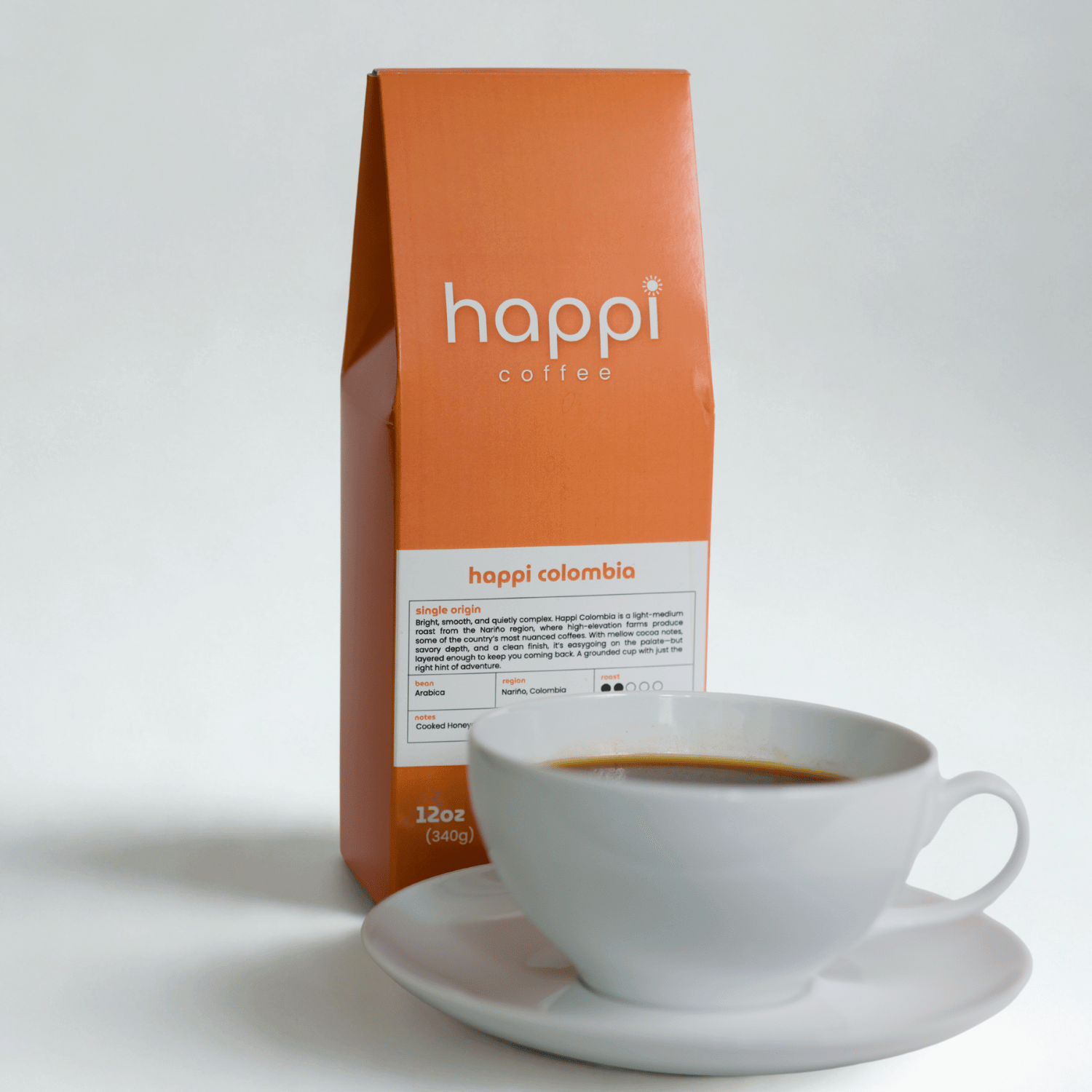 View of 12 oz bag of Happi Colombia next to a cup of coffee. Best Light Roast Coffee Beans with Bright Citrus, Cocoa Warmth & Smooth Body, Available Ground or Whole Bean